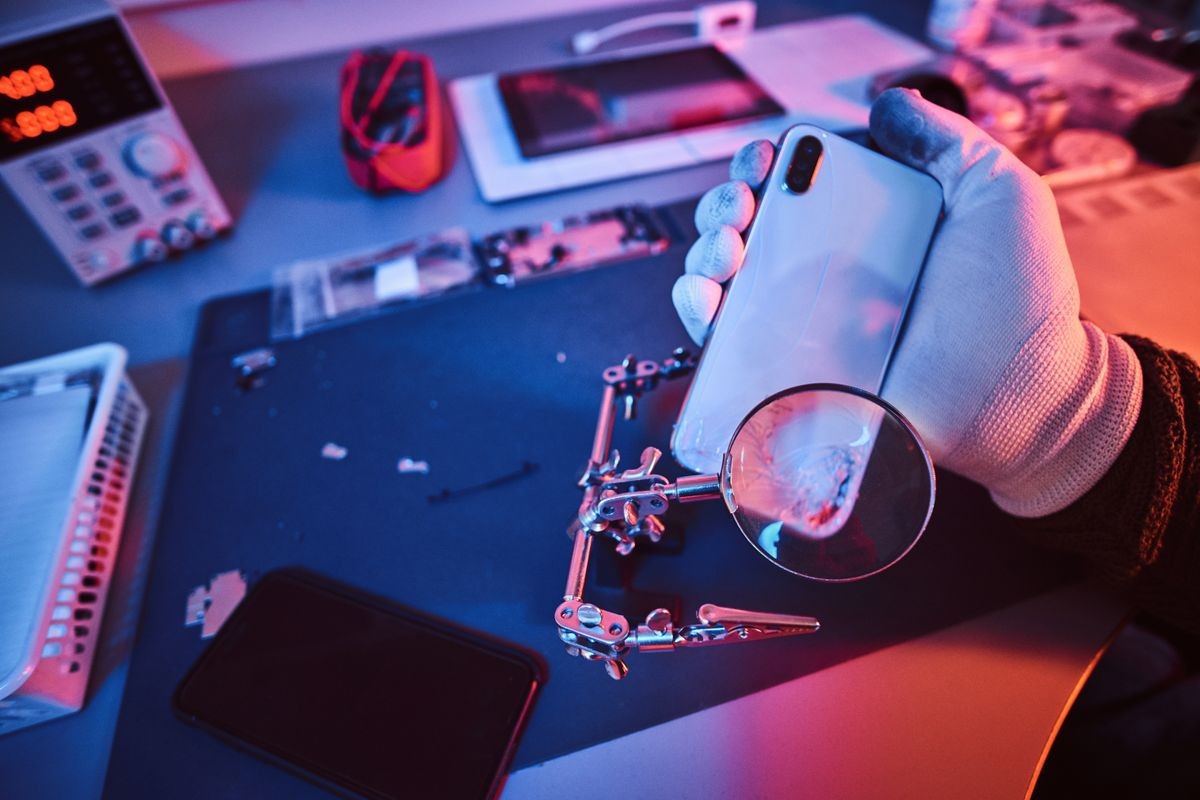 Electronic technician holds a modern smartphone with a broken body, carefully examines the damage using a magnifying glass sitting at the desk in the repair shop. Illumination with red and blue lights